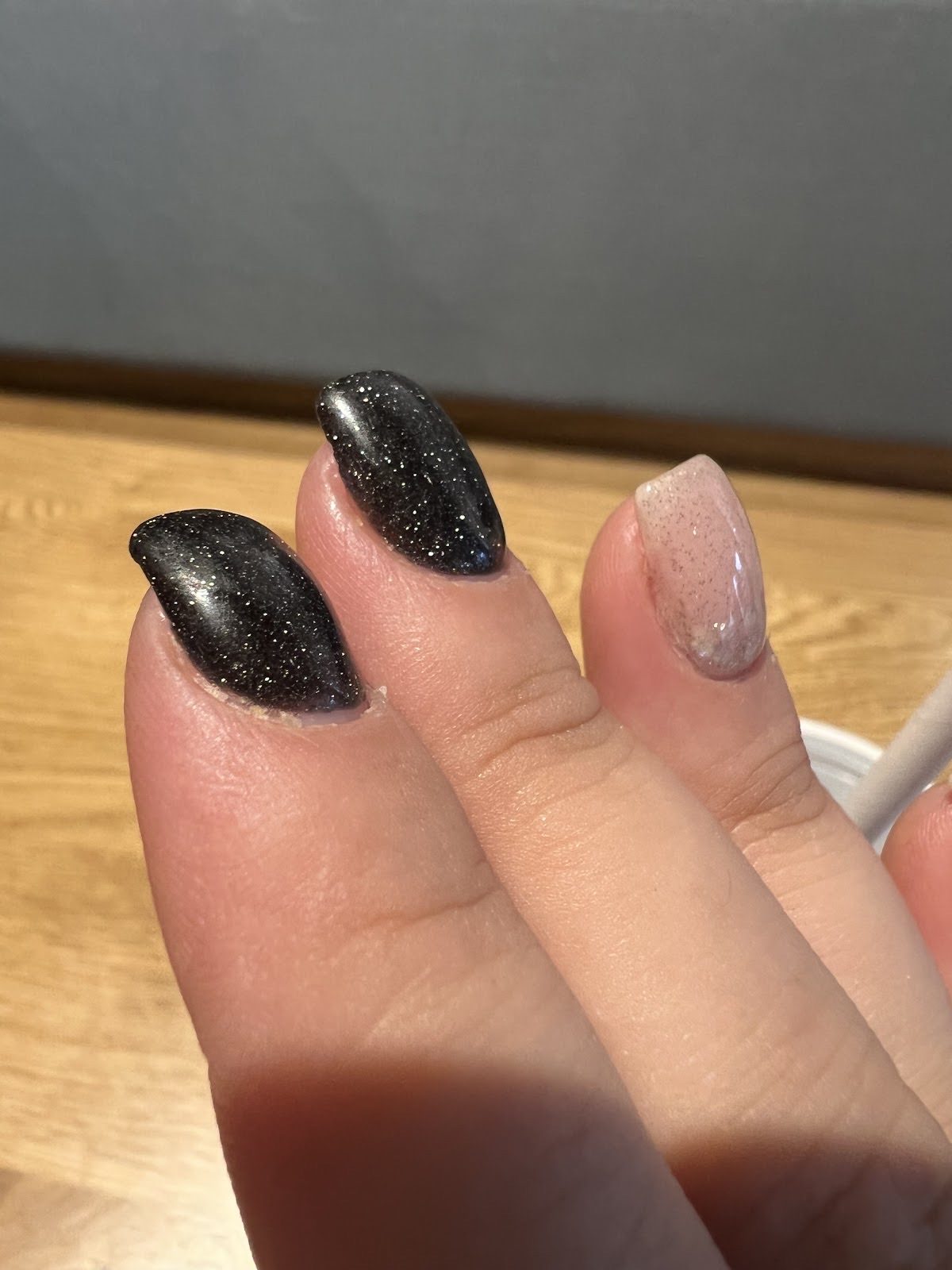 Hand over a wooden table with black glitter nails and a nude glitter accent nail.